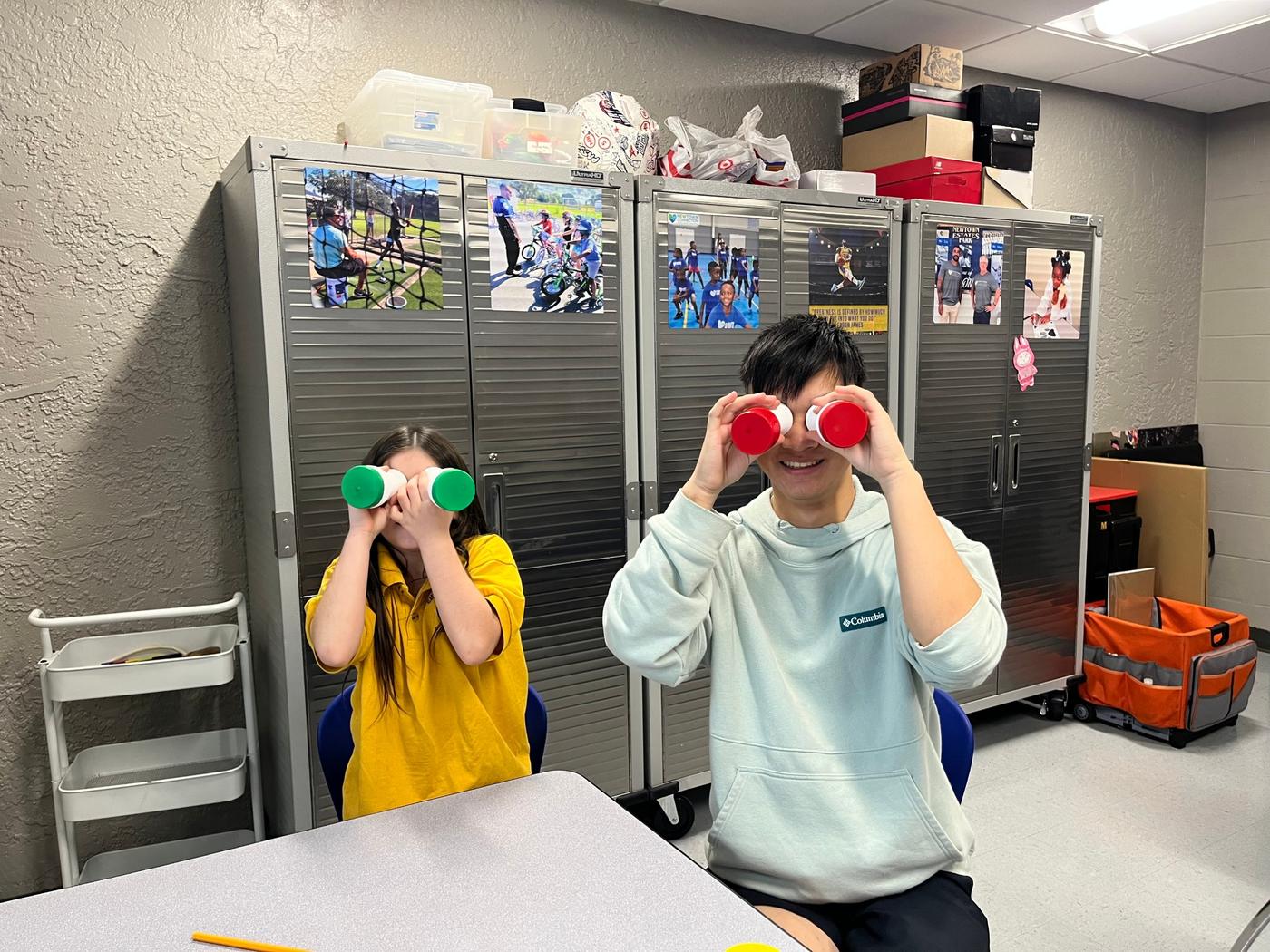 A volunteer and a student playfully hold their instruments up to their eyes like binoculars.