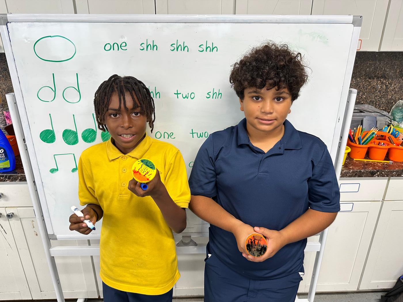 Two students hold up their decorated instruments in front of a whiteboard of rhythm notation.
