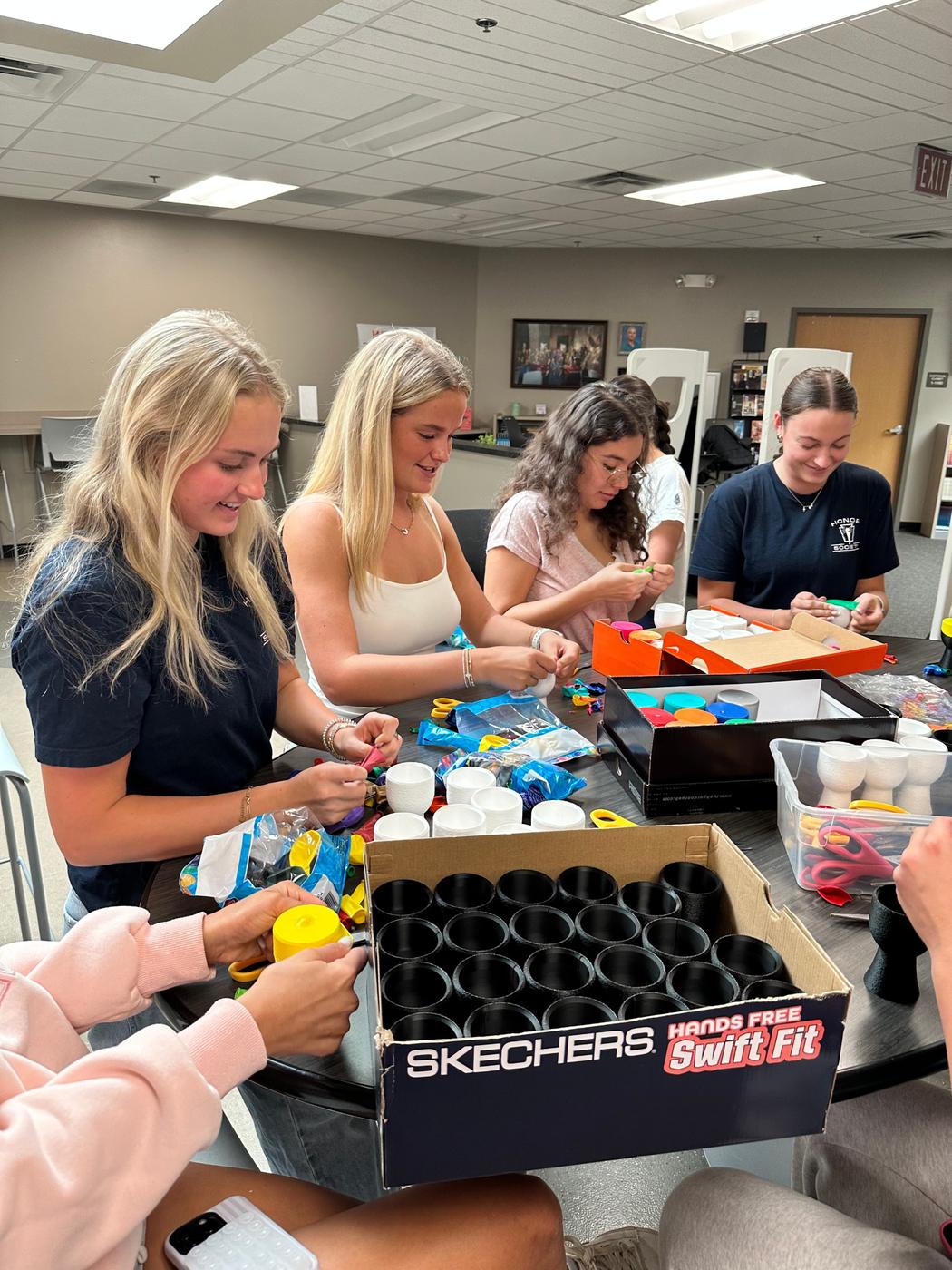 Volunteers assemble hand drums at a table, surrounded by balloons, scissors, and boxes of printed parts.
